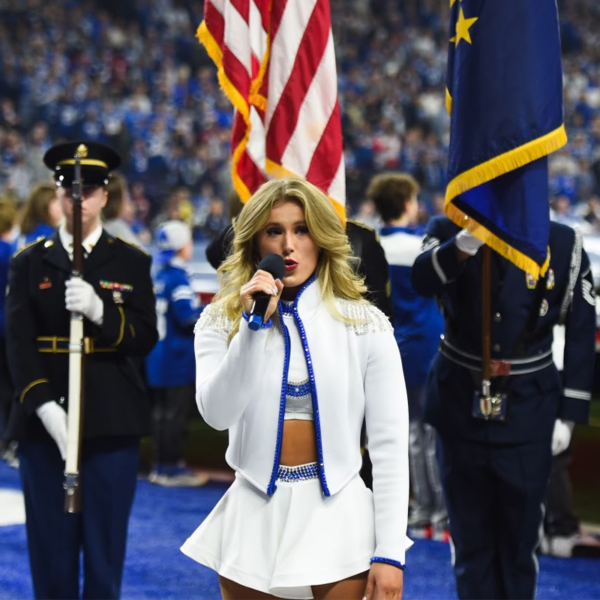 Woman with blonde hair and wearing cheerleader uniform holds a mic and is singing in front of soldiers in uniform and the American flag.