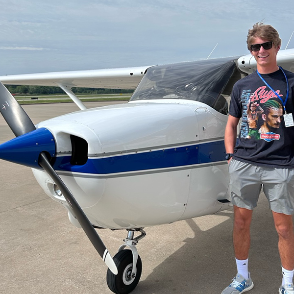 Young man in t-shirt, sunglasses, and cargo shorts stands in front of a small, two-seater plane. Behind the plane is a runway and open field.