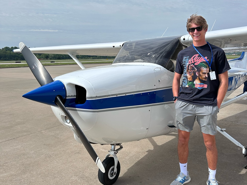 Young man in t-shirt, sunglasses, and cargo shorts stands in front of a small, two-seater plane. Behind the plane is a runway and open field.