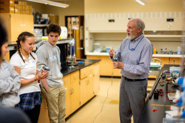 A teacher and three students stand in a science lab. The teacher is showing the students a device attached by a wire to another part held by a student.