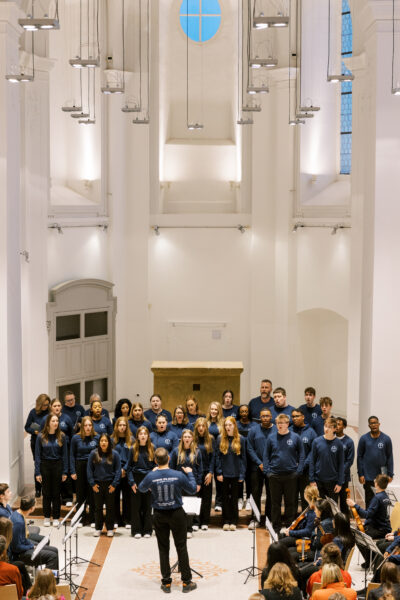 Choir students stand in front of an audience inside of a white church with vaulted ceilings.